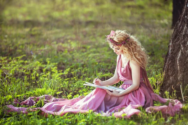 Girl in a fairy dress sitting under a tree in the woods and draws