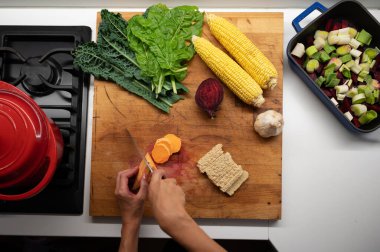 Renkli sebzelerle dolu bir tahta üzerinde patates kesen kadın ellerinin üst görüntüsü - mısır, pancar, lahana, sarımsak ve tempeh. 