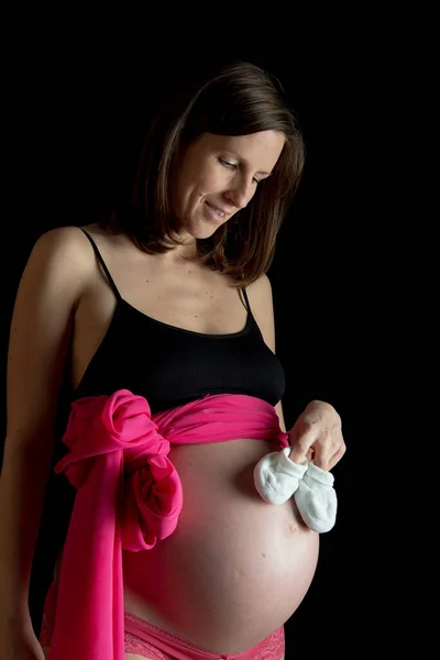 ピンクのリボンと赤ちゃんのスリッパで彼女の大きな腹を愛して幸せな若い妊婦さん 黒を背景に — ストック写真