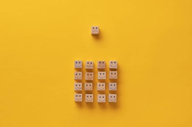 Hüzünlü bir yüzle tahta blok, kavramsal bir imajla gülen yüzlü bir grubun üstünde. Sarı arkaplan.