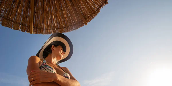 Low angle view of a young woman in a bikini and sunhat standing under sun umbrella on a beautiful clear summer day.