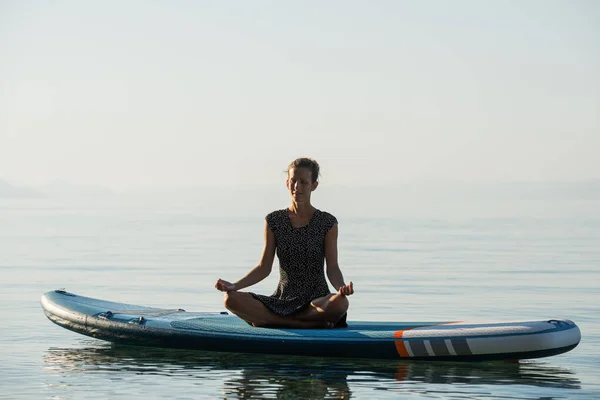 Peaceful young woman with a serene expression on her face meditating in lotus position on a sup board floating on water.