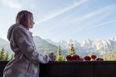 Pembe saçlı, sıcacık kazaklı genç bir kadın ahşap bir balkon çitine yaslanıyor, elinde bir fincan kahve tutuyor ve dağların güzel manzarasının tadını çıkarıyor..