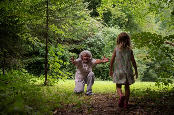 おじいちゃんは美しい緑の草原の端でひざまずいて彼の孫娘を迎えるためにオープン腕で — ストック写真