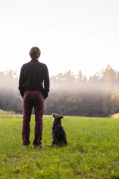 Young man standing in the middle of green meadow with his black — Stock Photo, Image