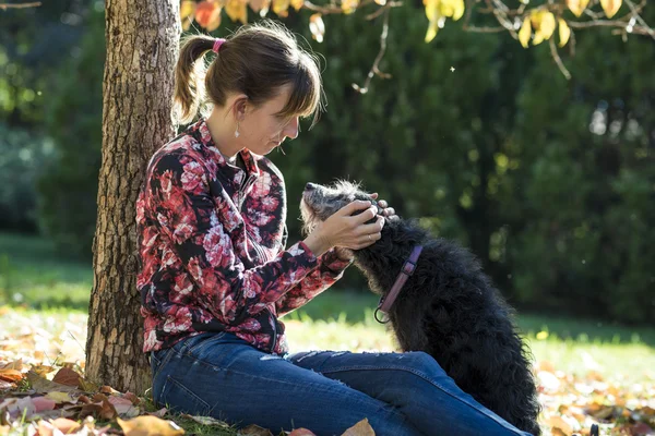 Jovem mulher sentada sob uma árvore de outono abraçando seu cão preto — Fotografia de Stock
