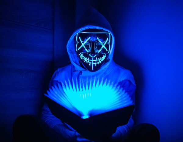 Man in glowing mask sitting in the corner with book-shaped lamp 
