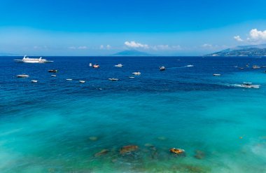 Capri Adası 'nda küçük bir sahilin yanında mavi masmavi bir sahil ve denizde birkaç tekne var.
