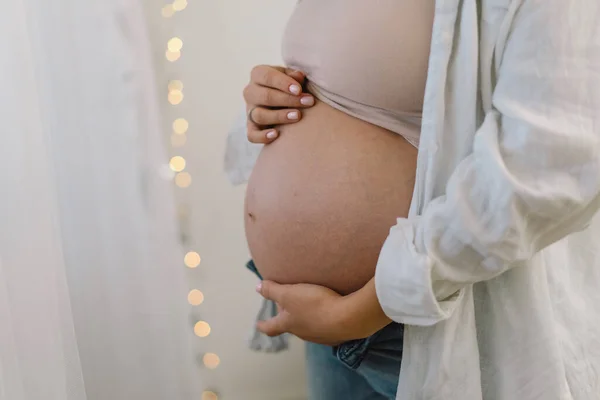 Atractiva mujer embarazada está de pie junto a la ventana y sosteniendo su vientre. Últimos meses de embarazo — Foto de Stock