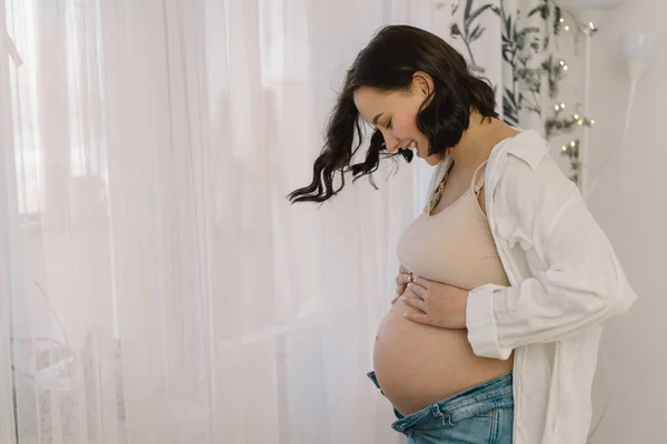 Atractiva mujer embarazada está de pie junto a la ventana y sosteniendo su vientre. Últimos meses de embarazo — Foto de Stock