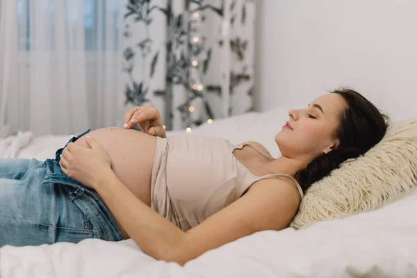 Atractiva mujer embarazada está sentada en la cama y sosteniendo su vientre. Concepto de embarazo. — Foto de Stock