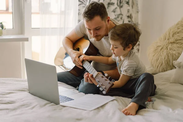 Father and son learn to play the acoustic guitar in an online lesson. Free time to spend with my son at home, teaching him to play the guitar. Happy Father\'s day