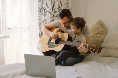 Baba ve oğul online bir derste akustik gitar çalmayı öğrenirler. Oğlumla evde vakit geçirmek, gitar çalmayı öğretmek için. Babalar günün kutlu olsun.