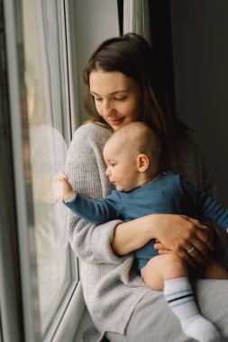 Anne, bebek oğluyla pencerenin yanında oynuyor. Mutlu bebek ve anne. Anneler gününde karantina sırasında izolasyon