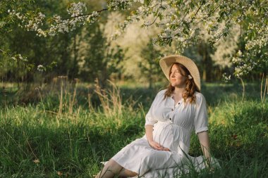 Hamile bir kadının bahar portresi. Beyaz elbiseli güzel hamile bir kadın bahar bahçesinde yürüyor..