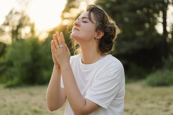 Wanita menutup matanya, berdoa di lapangan saat matahari terbenam yang indah. Tangan dilipat dalam konsep doa untuk iman — Stok Foto