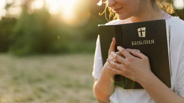 Christian woman holds bible in her hands. Reading the Holy Bible in a field during beautiful sunset.