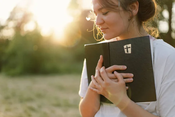 La donna cristiana tiene la Bibbia tra le mani. Leggere la Sacra Bibbia in un campo durante il bel tramonto. Foto Stock Royalty Free