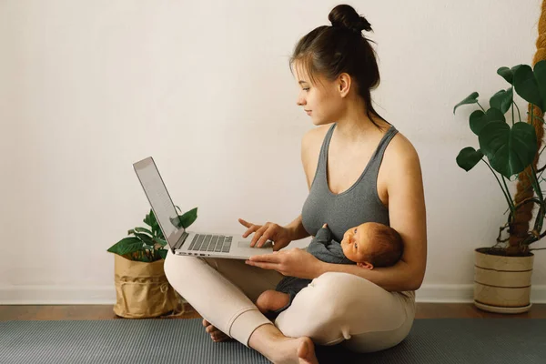 Mother holds her newborn baby son and works at the computer at home.