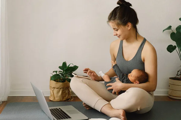 Mother holds her newborn baby son and works at the computer at home.