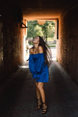 Mavi elbiseli mutlu genç bir kadın sokakta kameraya bakıp gülüyor. Şehir arkaplanı