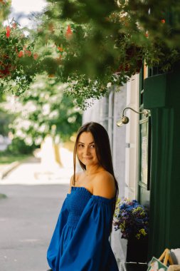 Mavi elbiseli mutlu genç bir kadın sokakta kameraya bakıp gülüyor. Şehir arkaplanı