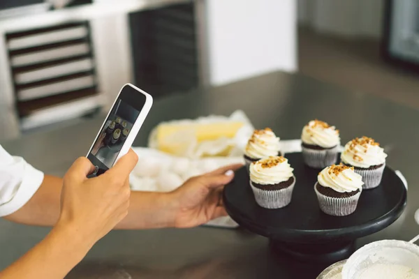 Chica pastelera fotografiando magdalenas para su blog. Chica hace una foto de cupcakes en un teléfono inteligente — Foto de Stock