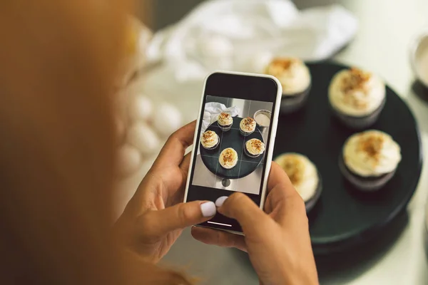 Chica pastelera fotografiando magdalenas para su blog. Chica hace una foto de cupcakes en un teléfono inteligente — Foto de Stock
