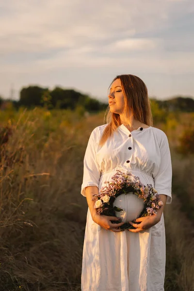 Retrato de una mujer embarazada. Una hermosa joven embarazada en un vestido blanco camina en el campo. — Foto de Stock