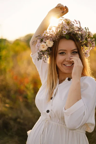 Beautiful woman in a wreath of wild flowers. Standing back in the flower field, hands to the side — Stock Photo, Image