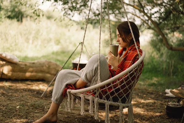 สาวสวยห่อในลายสก๊อตสีแดงดื่มชาในเก้าอี้แขวนที่อบอุ่นกลางแจ้ง การเดินทางผจญภัย — ภาพถ่ายสต็อก
