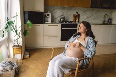 Hamile bir kadın şık bir mutfakta elinde bir fincan çayla duruyor ve huzurlu bir ortamda yaklaşan anneliği düşünüyor.