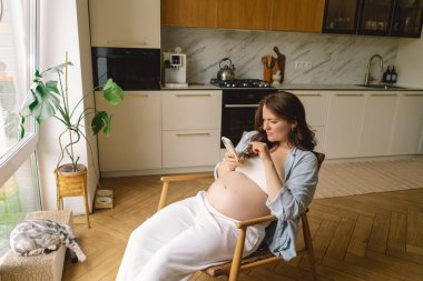 Hamile kadın çağdaş bir mutfakta duruyor, akıllı telefonuna odaklanmış. Doğal ışık mekanı aydınlatır, rahat ve şık bir ortam sunar..