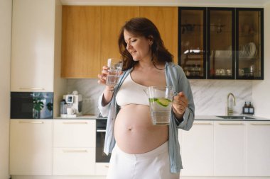 Günümüz mutfağında, neşeli bir suratı olan hamile bir kadın bir sürahi dolusu su ve bardak tutuyor. Parlak ışık, o su içmekten zevk alırken onun ışıl ışıl gülümsemesini arttırır..