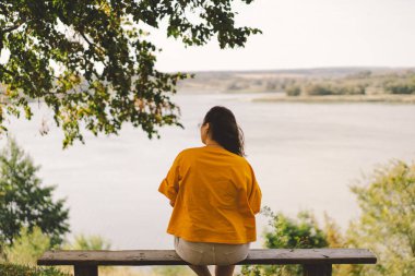 Sakin nehir kıyısında, sarı tişörtlü bir kadın bir bankta oturur, suyu ve uzak tepeleri seyreder, etrafındaki sakin manzarayı emer..