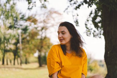 Parlak sarı bluzlu bir kadın, sonbaharda güneşin sıcaklığından ve doğanın güzelliğinden zevk alırken mutlu bir şekilde parkta yürür..