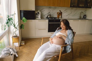 Sıcak bir mutfakta hamile bir kadın hamile karnını nazikçe tutuyor. Huzurlu bir ortamda yaklaşan anneliği yansıtıyor.