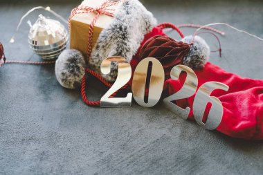 2026 yılı için parlak sayıların yanına ılık bir içecek konur, şenlikli süsler ve yumuşak dekorasyonlarla çevrili, rahat bir kış atmosferi yaratır. Mutlu Noeller Mutlu yıllar 2026