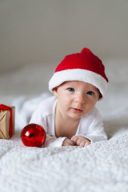 A baby wearing a red Santa hat lays on a cozy white blanket surrounded by festive decorations. A small gift box and shiny ornaments add to the cheerful holiday spirit.
