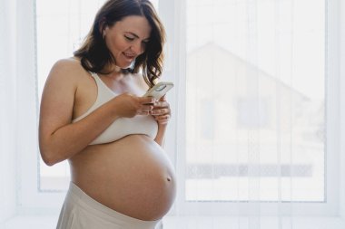 A pregnant woman stands by a bright window, smiling as she uses her phone. She is dressed in comfortable clothing, enjoying a serene moment in her home. This moment captures the beauty of pregnancy.