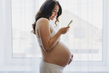 A pregnant woman stands by a bright window, smiling as she uses her phone. She is dressed in comfortable clothing, enjoying a serene moment in her home. This moment captures the beauty of pregnancy.