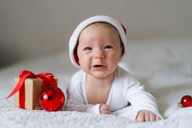 A baby wearing a red Santa hat lays on a cozy white blanket surrounded by festive decorations. A small gift box and shiny ornaments add to the cheerful holiday spirit.