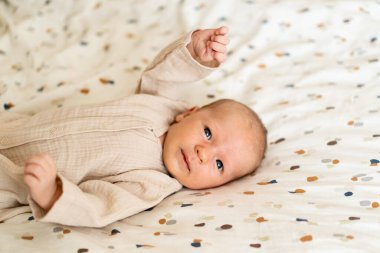 Yumuşak, renkli bir battaniyenin üzerine uzanmış yeni doğmuş bir bebek meraklı bir şekilde geniş gözlerle izleyiciye bakıyor. Sıcak atmosfer, bebeklerin nazik ve masum özelliklerini vurgular..