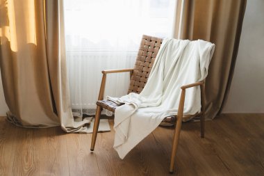 Basit bir tahta koltuk, ılık güneş ışığıyla yıkanmış yumuşak beyaz bir battaniye tutar. Perdeler pencereyi çerçeveler, sakin ve davetkar bir atmosfer yaratır..