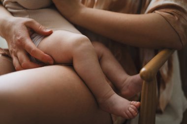 Tahta bir sandalyede oturan bir anne, bebeğini sıcak ve yumuşak bir odada nazikçe emziriyor. Güneş ışığı perdelerin arasından süzülüp barışçıl bir atmosfer yaratır..