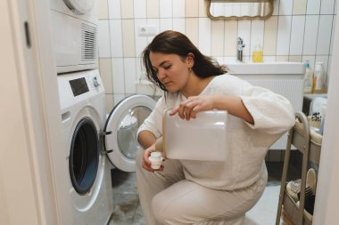 Çamaşır odasında çömelmiş çamaşır makinesine deterjan döken bir kadın. O, göreve odaklandı. Odada çamaşır ve temizlik malzemeleri var..