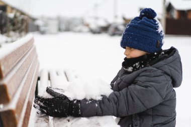 Mutlu çocuk dışarıda kışın tadını çıkarıyor. Sıcak kışlık giysiler ve örgü şapka giyen, soğuk bir kar gününde karda oynayan gülümseyen çocuk. Doğal ışık yaşam tarzı portresi ve çocukluk mutluluğu.