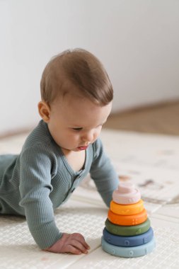 Yumuşak bir oyun hasırının üzerinde karnının üzerine uzanmış duyusal bir oyuncakla oynayan sevimli bir bebek. Bebek, oyuncağı keşfederken odaklanmış ve meraklı, erken öğrenme ve motor beceri geliştirmeyi destekliyor.