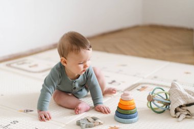 Yumuşak bir oyun hasırının üzerinde karnının üzerine uzanmış duyusal bir oyuncakla oynayan sevimli bir bebek. Bebek, oyuncağı keşfederken odaklanmış ve meraklı, erken öğrenme ve motor beceri geliştirmeyi destekliyor.