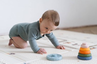 Yumuşak bir oyun hasırının üzerinde karnının üzerine uzanmış duyusal bir oyuncakla oynayan sevimli bir bebek. Bebek, oyuncağı keşfederken odaklanmış ve meraklı, erken öğrenme ve motor beceri geliştirmeyi destekliyor.